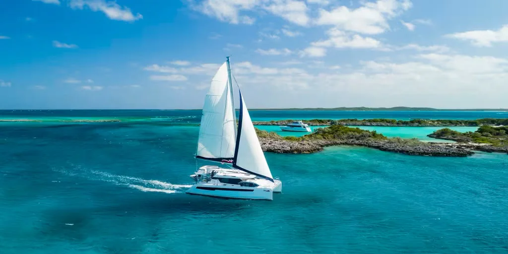A catamaran under full sail through turquoise water past a low Bahamian cay