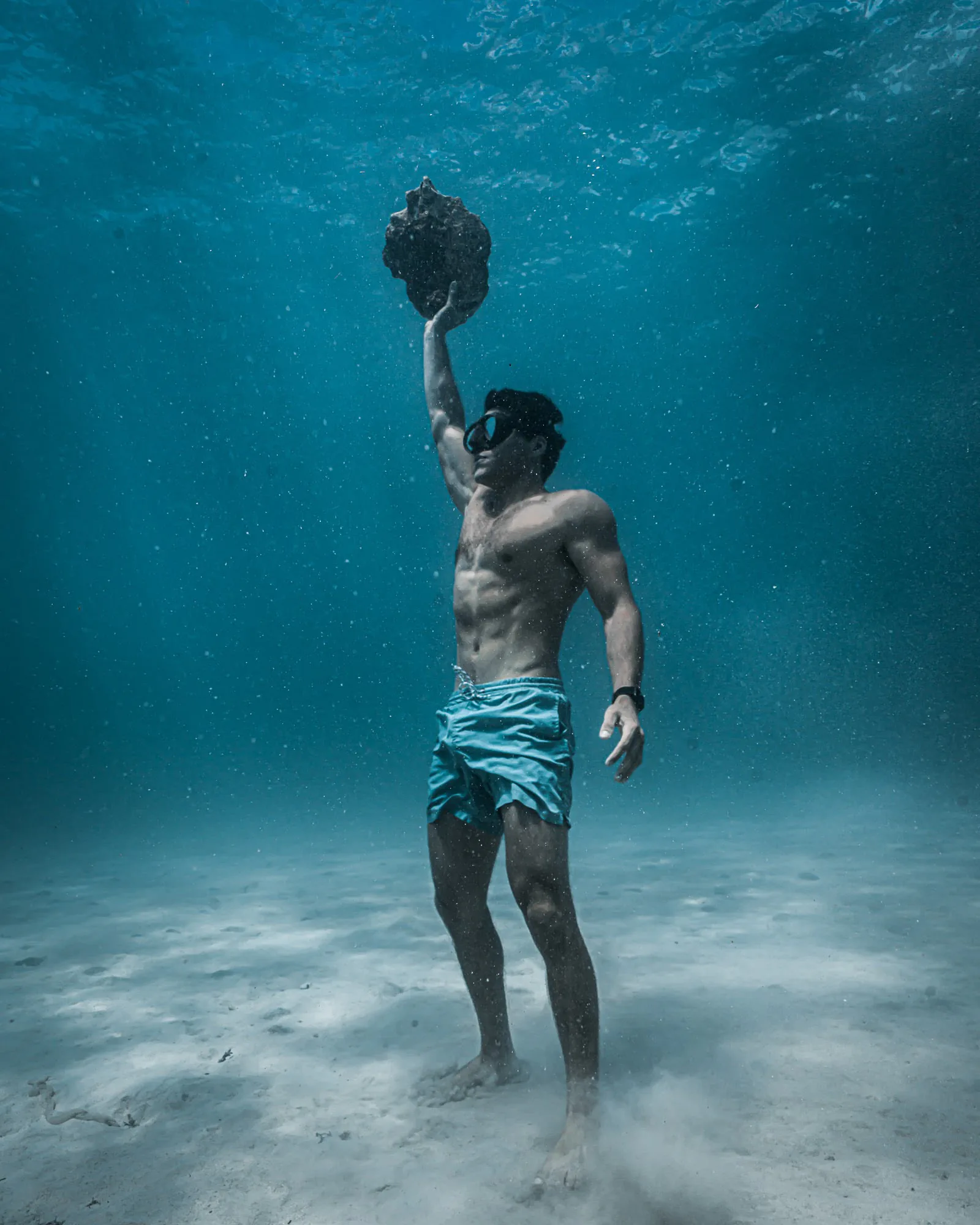 A freediver standing on the white sand bottom holding a conch shell overhead, sunlight scattering above